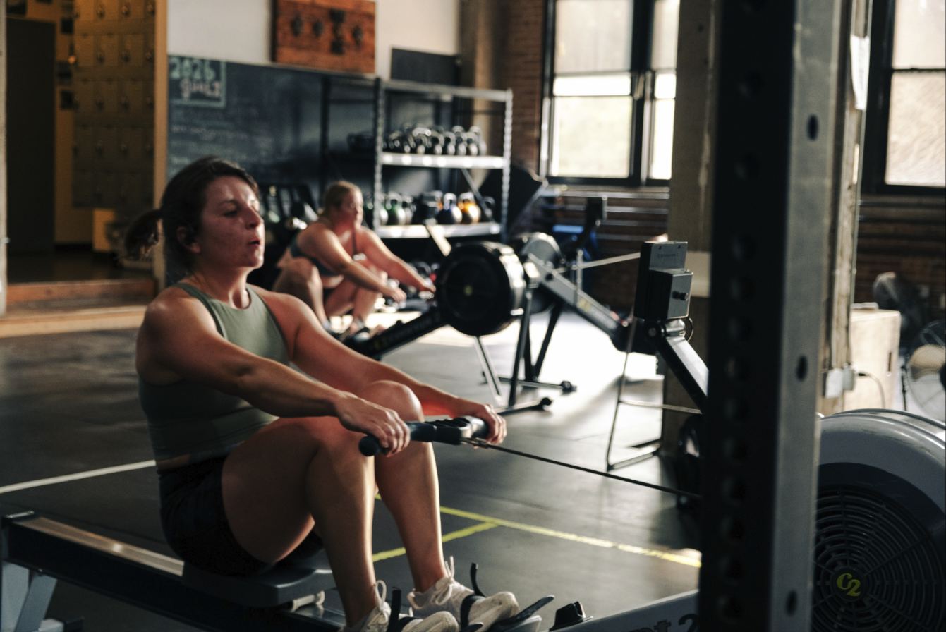 Two members rowing on Concept2 rowers under the gym's industrial windows