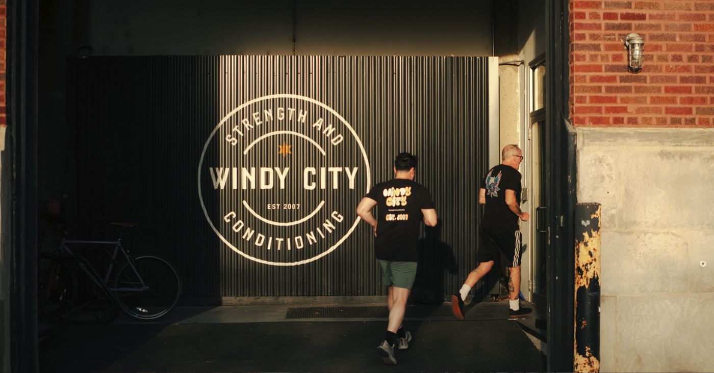 Two members jogging past the Windy City Strength and Conditioning sign at the gym entrance at golden hour
